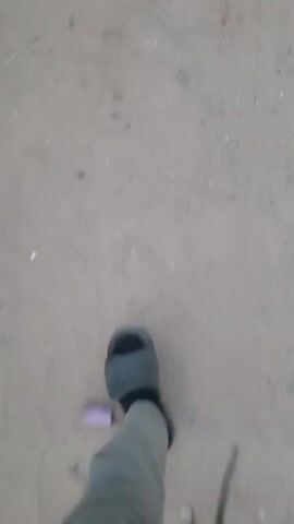 The video shows a first-person perspective of someone walking on a sandy, uneven surface. Debris such as small rocks and what appear to be pieces of plastic are scattered across the ground. In the foreground, the person's feet are visible, wearing dark-colored shoes. The camera is moving, indicating the person is in motion, and the scene is somewhat blurry due to the movement. There are no other people or significant objects clearly visible in the frame besides the ground and the person's feet.