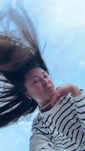 The video shows a young woman with long, dark hair against a bright blue sky. Her hair is being tossed around by the wind, creating a dynamic effect. She is wearing a white and black striped long-sleeved shirt and a pearl necklace. The camera angle is from below, looking up at her. The woman makes various facial expressions and gestures, including smiling, puckering her lips, and waving. The video appears to be a selfie taken outdoors on a windy day.
