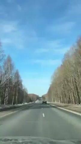 The video shows a view from a car driving on an asphalt road. The road is straight and stretches into the distance, with trees lining both sides. A truck is visible ahead on the road, moving in the same direction. The sky is bright blue with sparse clouds. The trees are mostly bare, suggesting it may be late fall or early spring. The road has white lane markings.