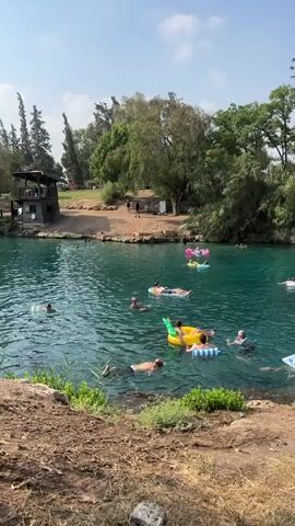 A group of people are enjoying a sunny day swimming and floating on inflatables in a clear blue body of water. The water is surrounded by lush green trees and foliage. In the background, there is a small building and a dirt path. Some individuals are swimming in the water, while others are relaxing on inflatable mattresses and toys. The overall atmosphere appears to be one of leisure and recreation.