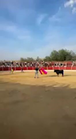 This video captures a bullfighting event in a traditional bullring, likely in San José del Valle, Spain, given the provided context. The scene is set under a clear blue sky with some scattered white clouds, suggesting it's daytime, possibly late afternoon due to the angle of the sunlight creating shadows on the sandy arena floor.

In the center of the dusty bullring, several individuals dressed in bullfighting attire are present. One matador is prominently featured, holding a vibrant pink and yellow cape, engaging with a black bull. Another matador is visible in the background, also maneuvering around the bull. The bull itself is dark-colored and appears active, moving around the arena.

The stands surrounding the arena are filled with spectators, though their faces and individual details are blurred. They are seated in rows, likely on benches or seats, watching the action unfold in the ring. The overall atmosphere appears to be that of an ongoing spectacle or performance. No discernible text is visible within the video frame.