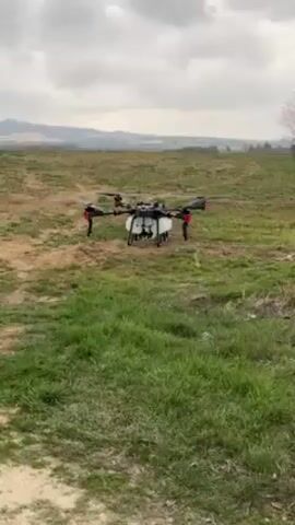 The video depicts a large, multi-rotor drone taking off from a grassy field. The drone is black and white with red accents and has a tank mounted underneath. The background features rolling hills and a cloudy sky. The camera follows the drone as it rises and flies into the distance. The drone is the main subject of the video and its upward movement is the primary action.