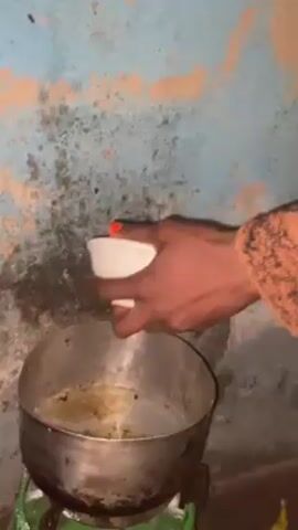 In this video, an individual wearing a striped sweater pours a light yellow liquid from a white ceramic cup into a metal pot containing hot oil. The pot sits on a small green gas stove. The liquid sizzles upon contact with the oil. The background features a worn wall painted in light blue with patches of brown. The person's hands are dark-skinned with one fingernail painted red.