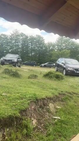The video shows a grassy field with several cars parked on it, against a backdrop of trees and a partly cloudy sky. The view is framed by the edge of a wooden structure, likely a porch or overhang, suggesting the video is taken from within a shelter of some kind. In the foreground, there is an eroded dirt bank. The cars include a dark-colored SUV on the left, a dark-colored compact car slightly behind it, a silver sedan in the center, and a dark-colored station wagon on the right. The overall impression is of a rural setting where people have gathered, possibly for an event or picnic.