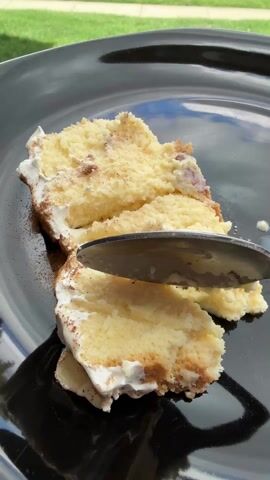 The video shows a slice of cake being cut with a spoon on a black plate, outdoors. The cake is a light yellow color with white frosting along one edge and some kind of darker sprinkles or topping.  The plate reflects the sky, suggesting it is a sunny day.  A green lawn is visible in the blurred background. An off-screen person uses a spoon to cut a piece from the slice, then cuts that piece into smaller bites.