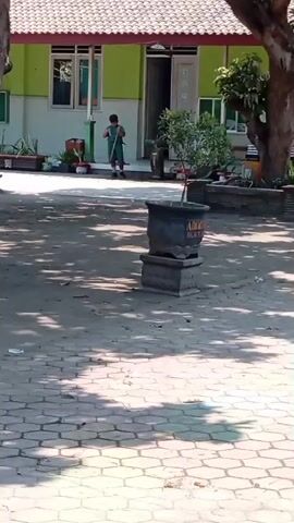 A person wearing a green uniform is sweeping a paved courtyard in front of a light green building. The building has a white roof with red trim, multiple windows, and a doorway. There are trees and potted plants in the courtyard. The person is holding a broom and appears to be moving it across the ground. The video is shot from a medium distance.