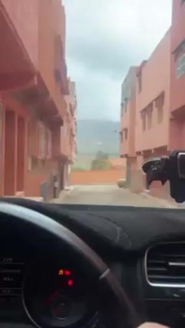 The video shows a view from inside a car, likely the driver's perspective. The car is on a narrow street lined with buildings painted in a reddish-orange color. The buildings are two or three stories high and have rectangular windows with shutters. The sky is overcast. In the distance, there are rolling hills or mountains. The dashboard of the car is visible, including the steering wheel, instrument cluster, and air vents. The windshield wipers are activated, clearing rain or dirt from the windshield. The dashboard displays digital information.