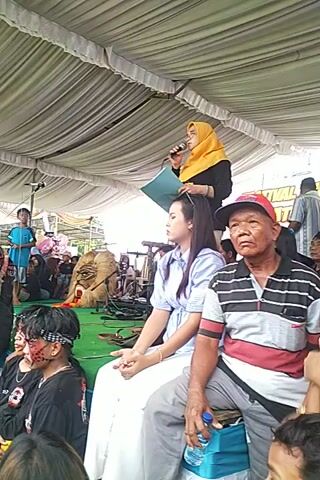 Here is a forensic description of the video:

The video shows a woman in a light blue top and dark pants interacting with a man on the ground who has dark makeup on his face. They appear to be on a stage or platform, covered with a green rug under a tent-like structure. Behind them, a woman in a yellow hijab stands holding a blue folder, seemingly giving a speech or announcement as she speaks into a microphone. Several other people are visible, including a man in a red cap and a striped shirt. The atmosphere is crowded, and the scene seems to be part of an event or performance taking place outdoors.