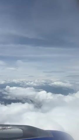 This is a vertical, POV video from inside an airplane cabin, looking out the window. The airplane's engine nacelle and wing are visible in the foreground, occupying the bottom of the frame. Above this, the image transitions to a view of the sky. Numerous white, puffy clouds are spread out below, creating a layered effect. Above the cloud layer, a vast expanse of sky stretches to the horizon, filled with thinner, gray, streaky clouds. The overall lighting suggests daylight, possibly a cloudy day or a time when the sun is not directly visible. The camera is stationary throughout the short clip, capturing a static aerial landscape.