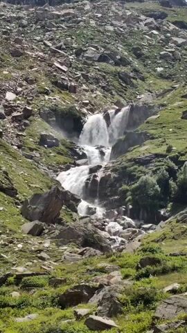 A vertical shot shows a cascading waterfall flowing down a rocky, grassy mountainside. The water tumbles over several ledges, creating a misty spray. The surrounding terrain is covered in rocks and patches of green vegetation. The sky above is partly cloudy.