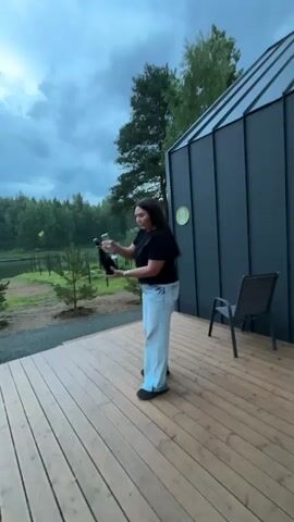 A woman stands on a wooden deck, holding a bottle of champagne and a glass. She is wearing a black t-shirt and light blue jeans. She pops the cork off the bottle, which flies into the air. She then runs towards the camera, smiling. In the background, there is a modern house, trees, and a body of water. The sky is cloudy.
