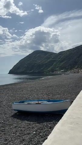 The video displays a serene coastal landscape in Lipari, Italy, on a bright, partly cloudy day. In the foreground, a small, white and blue boat rests on a dark, pebbly beach. The tranquil sea stretches out to the left, reflecting the soft light. In the background, lush, green mountains rise majestically from the water, their slopes covered in vegetation. Along the distant shoreline, a scattering of white and pastel-colored buildings suggests a small village or town nestled at the base of the mountains. The sky is a light blue, adorned with fluffy white clouds, and the overall atmosphere is one of calm and natural beauty. No people are visible, and no text is present in the video.