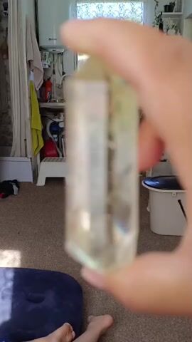 A close-up shot shows a hand holding a clear, faceted crystal with a light yellow hue. The crystal is positioned vertically, with its top pointed end facing upwards. The background is blurred, but suggests an indoor environment with shelves and possibly some clothing hanging. Sunlight appears to be entering from the left, casting shadows and illuminating the crystal. The crystal has some internal inclusions or cloudiness, visible as white wispy patterns. The hand holding the crystal appears to be an adult's, with visible fingernails.