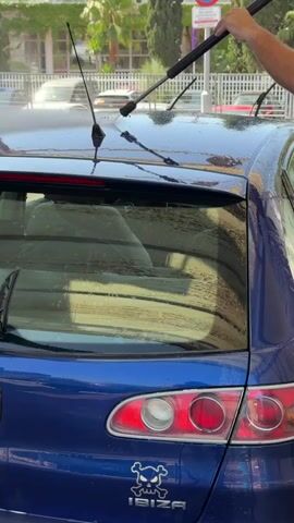 The video shows a person pressure washing the rear of a blue car. The car has a rear windshield wiper, a spoiler, and a sticker of a skull and crossbones with "IBIZA" written underneath. The car is parked outdoors, likely at a car wash, with a building and palm trees visible in the background. The sun is shining, indicating it is daytime, and the weather appears to be clear. The pressure washer is being held by a person whose arm and hand are visible in the top right corner of the frame. The water spray is creating droplets on the car's surface. The overall scene depicts a car wash activity.