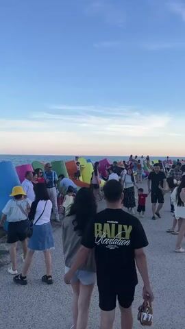 A group of people are gathered on a walkway next to the ocean.  Large, colorful, concrete structures, possibly breakwaters, line the edge of the water. Some people are standing, some are sitting, and some are walking.  Several children are visible, some interacting with the structures and others with adults. The sky is clear and blue, suggesting it is daytime.