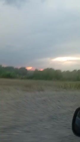 The video shows a blurry view of a landscape with trees and a dusty road. The sky is overcast with some pink and orange hues, suggesting either sunrise or sunset. The camera appears to be moving, possibly from inside a vehicle, as indicated by the motion blur. There are no people or discernible objects other than the natural environment.