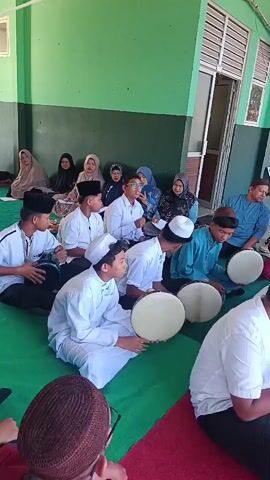 A group of young males, dressed in white Islamic attire and caps, are seated on the floor, playing frame drums. Several adults, also in Islamic clothing, are observing the performance. The setting appears to be an outdoor covered area with green walls and a tiled floor. Sunlight streams into the scene.