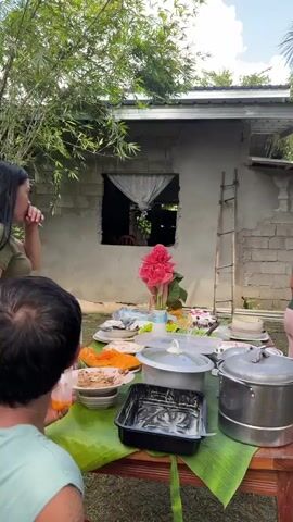 A group of people is gathered around a wooden table set outdoors. The table is covered with banana leaves and holds several dishes, including a pot of food, a cake, and a bowl of fruit. A woman in a green shirt is seen moving food from a tray to a plate. Another woman in a striped shirt and pink pants is standing at the end of the table. A man in a light green shirt is seated at the table, facing away from the camera. In the background, there is a house under construction and various trees and foliage.