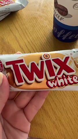 Here is a detailed description of the video:

A hand holds a Twix White candy bar in front of a wooden table. The candy bar is wrapped in a white wrapper with red lettering that reads "Twix White." The wrapper also has an image of the candy bar and a small symbol. 

In the background, there is a white paper cup with a blue design and the word "coffee" written on it. There is also another wrapped candy bar visible in the background.