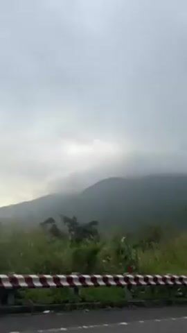The video shows a roadside view with a mountain range in the background under a cloudy sky.  A red and white barrier lines the side of the road. A cyclist wearing a helmet and blue clothing rides on the road.
