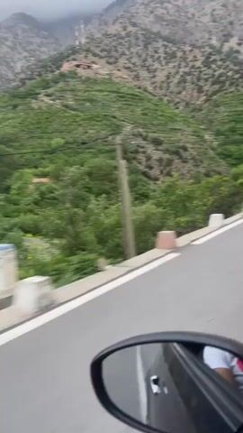 The video shows a drive along a winding mountain road, likely from the perspective of a vehicle's passenger side, with part of a car mirror visible in the bottom right corner. The road has a white solid line on the left and a white broken line on the right, bordered by low, white and red concrete guardrail blocks.

The surrounding landscape is mountainous and rugged. To the right of the road, a steep mountain slope rises, covered in light-colored rocks and sparse, low-lying green vegetation. A power pole stands by the roadside, and further along, a stone wall and some satellite dishes are visible on the side of the mountain.

To the left and below the road, a valley is filled with lush, dense green trees and bushes, suggesting a river or more fertile ground at the base of the mountains. Several trees are scattered along the roadside, some appearing to sway in the wind. The sky is overcast and gray. The overall setting is a scenic, mountainous region in Asgaour, Morocco. No people are visible.