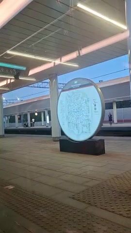 A circular digital display board stands on a train platform. The board illuminates with a map of train routes. A person is standing on the platform to the right of the board, looking away from the camera. Another person is walking on the platform to the left of the board. The platform is covered by a modern structure with bright lighting. The sky is visible through the structure.