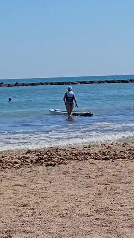 A person wearing a life vest and a hat stands in the shallow water next to a paddleboard. The person is facing away from the camera and appears to be getting ready to paddleboard. The sun is shining brightly, indicating it is daytime. The water is a clear blue and there are small waves breaking on the sandy beach in the foreground. In the distance, a breakwater made of rocks stretches across the water. Another person can be seen in the water further out. The scene is a beach in Deveses, Spain, on a clear and sunny day.
