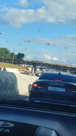 A dark-colored Kia Optima is shown from the rear on a highway. The car is in traffic, with other vehicles visible ahead. A bridge or overpass is under construction in the background. The sky is partly cloudy. The license plate of the Kia Optima is visible.