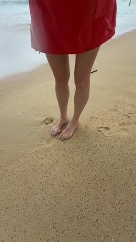 A person wearing a red skirt and bare feet stands on a wet sandy beach. The camera then pans to show the person holding a clear cup with a dark liquid and a blue straw. The camera then tilts to show the ocean waves, the sky with clouds, and a distant shoreline with trees.