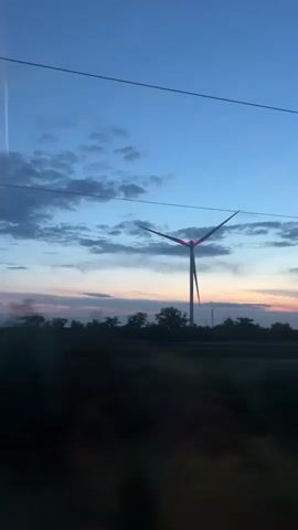 The video shows a view from a moving vehicle looking out at a landscape at dusk. A tall wind turbine with its blades spinning is visible in the middle of the frame, silhouetted against a blue and pink sky. Trees and other vegetation form a dark, blurry band across the lower part of the image. The motion of the vehicle causes the landscape to blur, especially in the foreground. There are power lines visible horizontally across the top of the frame.