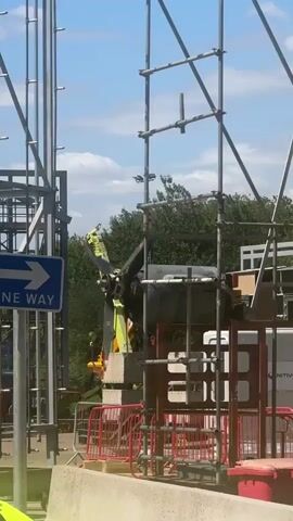 The video captures a construction scene with scaffolding and heavy machinery. A yellow crane with a large black object, possibly a rotor or blade, is positioned among the scaffolding. A blue and white "ONE WAY" sign is visible on the left. Construction barriers and a red bin are in the foreground, with trees and a blue sky in the background.