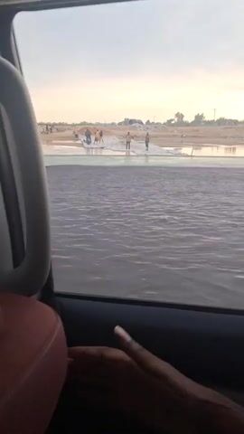 A person is recording from the inside of a vehicle as it is traveling on a road. Through the window, a scene of a body of water is visible, and a group of people are standing on what appears to be a sandbank or a land formation slightly above the water level. The group consists of about six individuals. Some are partially submerged in the water. The background beyond the people includes what seems to be piles of materials, possibly sand or construction related items, as well as some sparse vegetation. The overall atmosphere is one of a natural environment where people are engaged in some activity. The light suggests it could be either early morning or late afternoon.