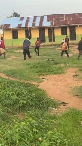 A group of young males are playing soccer on a dirt and grass field in front of a yellow and green building with a corrugated metal roof. One person is kicking the ball, and several others are positioned around the field, some watching and some actively participating in the game. The environment appears to be rural or semi-urban, with some vegetation in the foreground.