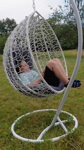 A young person is shown relaxing in a hanging egg chair outdoors. The chair is white and suspended from a white stand. The young person is wearing a light blue t-shirt, black shorts, and striped socks. They are lying down with their head resting on a dark gray cushion, and their legs are crossed. The chair is gently swaying. The background consists of green grass and trees. The sky appears to be overcast.