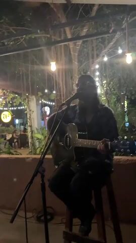 A man with long, dark hair and glasses, wearing a patterned shirt and dark pants, is seated on a stool, performing live music. He is playing an acoustic guitar, identifiable as a "Granada" from its headstock, and singing into a microphone positioned on a stand. He appears focused on his performance.

The setting is an outdoor or semi-outdoor venue, likely a cafe or restaurant, during the evening or night. The space is warmly lit by numerous string lights strung overhead, creating a cozy and inviting atmosphere. A prominent feature in the background is a large tree with many aerial roots hanging down, adding to the natural and tropical ambiance.

In the blurred background, other patrons can be seen seated at tables, enjoying the evening. A distinctive "a2" logo is visible on a sign to the left. The overall scene suggests a relaxed and enjoyable live music event in a warm climate, consistent with a location like Arambol, India.