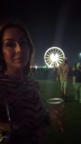 A young woman with shoulder-length brown hair drinks from a clear plastic cup with a straw, smiling at the camera. She is wearing a brown and black animal-print top and necklaces. In the background, there is a crowd of people, a Ferris wheel, and a tower illuminated with rainbow lights. The scene appears to be an outdoor festival at night.