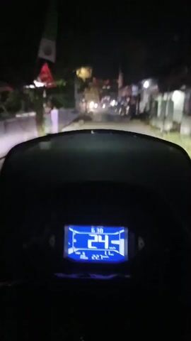 This is a first-person perspective video shot at night from a moving vehicle, likely a motorcycle, given the dashboard display. The dashboard shows a digital speedometer with the speed fluctuating between 24 and 25. The environment appears to be a residential street with buildings on either side. Streetlights and lights from the buildings illuminate the scene. The vehicle's headlights are on, casting a beam of light forward. The road surface appears to be wet, suggesting recent rain. The video is shaky, consistent with being recorded from a moving vehicle.