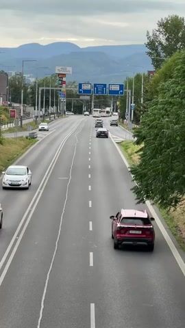 A vertical shot looks down on a multi-lane road with cars driving in the distance. The sky is overcast and a range of green mountains can be seen in the background. The road is paved and has white dashed lines marking the lanes. There are streetlights and signs along the side of the road, as well as trees and buildings. The cars are moving at a moderate speed. The overall scene suggests a suburban or urban environment during the day.