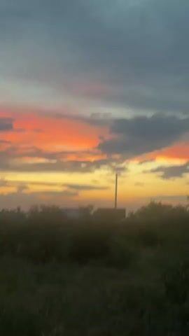 The video shows a time-lapse of a sunset with clouds. The sky is filled with colors ranging from gray to yellow and orange. In the foreground, there are trees and bushes. There is also a utility pole and what appears to be a small building. The camera pans slowly from left to right.