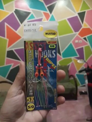 Here is a concise description of the video:

The video shows a hand holding a package of fishing lures. The lures are Mustad "StripeL" brand, saltwater big game lures with orange beads and colorful skirts. The package indicates the hook size is 5/0. The background is a wall with a geometric pattern of triangles in various colors.