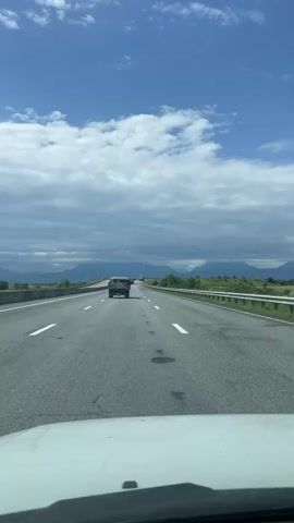 The video shows a first-person perspective from inside a vehicle driving on a highway. The road ahead has white lane markings and a guardrail on the right side. There is another vehicle, a dark SUV, driving in the same direction in the lane ahead. The sky is blue with scattered white clouds. In the distance, there are mountains and trees. The front of the dashboard of the vehicle the video is being filmed from is visible at the bottom of the frame.