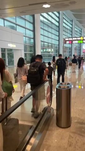 The video begins with a wide shot of an airport terminal. In the foreground, on the left, a person with long brown hair and a white top, carrying a green tote bag, is seen from the back, walking away from the camera and towards an escalator. To their right, a person in a light-colored outfit and a pink purse is also walking away. Further ahead, a man with a backpack is walking down the escalator. The escalator itself is made of metal with a glass panel. To the right of the escalator is a cylindrical trash can with two openings. In the background, the airport terminal is spacious and modern, with large windows and a high, curved ceiling. People are seen walking in various directions, some with luggage. There are signs with Chinese characters and directional arrows. The floor is a polished speckled material, reflecting the lights and the activity. The scene is well-lit, suggesting daytime or bright indoor lighting. The overall atmosphere is one of travel and movement within a public transit hub.