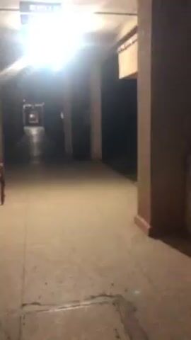 The video shows a nighttime view of a building exterior.  The camera moves along an open hallway with concrete columns and a dark ceiling. An open structure is visible across the hallway, allowing a view into what appears to be an interior room with windows.  There is a bright light fixture in the hallway ceiling. The footage is a bit shaky, suggesting it was filmed by someone walking.