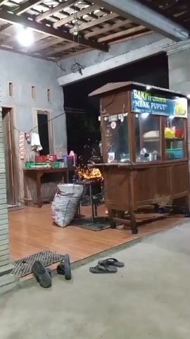 A wooden food stall with a glass display case is set up outdoors at night. The stall has a sign that reads "BAKMI JAWA MBAK PUPUT." Inside the stall, there are various containers of food and cooking utensils. In front of the stall, there is a grill with flames and sparks rising from it, suggesting food is being cooked. A large sack of what appears to be rice or flour is leaning against a metal structure near the grill. To the left of the stall, there is a wooden table with items on it, possibly ingredients or supplies. The ground is covered with wooden planks, and there are several pairs of sandals scattered around. The overall environment appears to be a humble outdoor setting, possibly a residential area or a small marketplace.