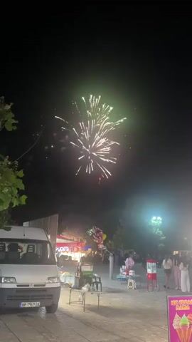 A nighttime scene shows fireworks exploding in the sky. In the foreground, a white van is parked on the left. In the middle ground, there are several stalls or carts with people gathered around them, along with some tables and chairs. Some of these stalls appear to be selling food or drinks, with one prominently displaying an "ICE CREAM" sign with colorful images of ice cream cones. A few people are standing and walking in the area. Trees are visible in the background, and artificial lights illuminate the scene. The overall atmosphere suggests a public gathering or event.