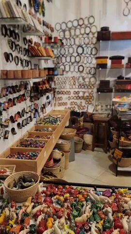 The video shows the interior of a gift shop full of handmade goods. Shelves, tables, and displays are filled with a variety of items, including wooden carvings, woven baskets, jewelry, and pottery. The shop is well-lit with white walls and a high ceiling. The camera slowly pans across the room, showcasing the extensive selection of merchandise. There are no people visible in the main shot.