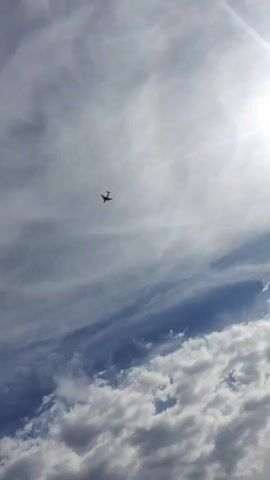 The video captures a wide shot of a cloudy sky with an airplane visible in the distance. The sun is shining brightly, creating a lens flare effect. The clouds are a mix of white and grey, with some appearing to be cumulus clouds. The airplane is a small silhouette against the vast sky, suggesting it is at a high altitude. The overall impression is one of a serene and expansive sky.