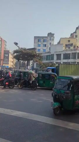 The video depicts a traffic intersection in an urban environment, likely in South Asia. The scene is viewed from the perspective of someone within or near a green three-wheeled vehicle. At the beginning of the video, there are several vehicles, including a truck loaded with boxes, motorcycles, and auto-rickshaws, waiting at a traffic light. People are visible on the motorcycles and near the truck. The traffic lights are visible and indicate red. The background includes commercial buildings and trees.

The camera angle shifts slightly, showing a broader view of the intersection. Traffic continues to wait at the intersection. The video ends by highlighting a large fountain in the middle of a roundabout. The fountain is surrounded by greenery and a low wall with murals or artwork. The sky is clear and blue. The video shows regular urban activity and focuses on traffic and the fountain as a landmark.