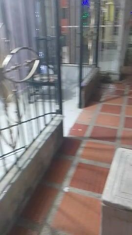 The video shows a metal gate with decorative elements from a low angle, looking upward. Beyond the gate, there's a glimpse of a room with a person visible in the reflection. The camera then pans down to show a brick floor, a white box on the floor, and a sandy or dirty ground area. The camera work is shaky and the footage is somewhat unclear.