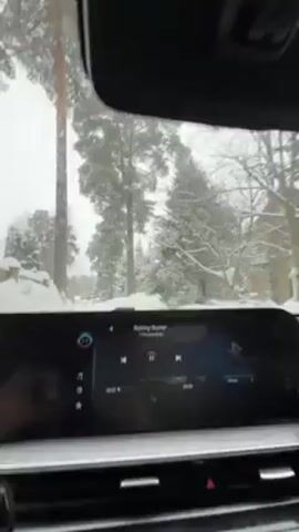 The video shows a view from inside a car driving in a snowy environment. The car's dashboard is visible, displaying a music track titled "Rolling Stoner." Outside the car, there are snow-covered trees and what appears to be a building in the distance, also covered in snow. Snowflakes are visible on the windshield, indicating active snowfall. The car is moving slowly through the snow-covered area.